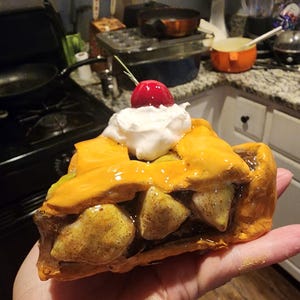 May include: A ceramic slice of apple pie with a golden-brown crust, visible apple chunks, a dollop of white whipped cream, and a bright red cherry on top. The piece has a glossy finish and is shaped to resemble a realistic dessert portion.