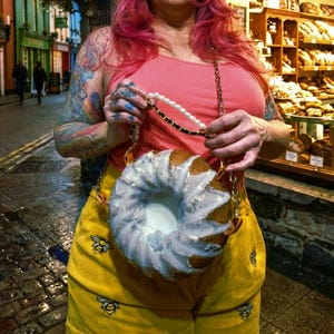 May include: A round, ring-shaped bag resembling a bundt cake with white icing, featuring a gold chain strap accented with white pearls. The bag is held against a coral-colored tank top and yellow pants embroidered with small black bee motifs.