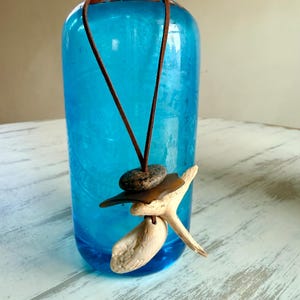 May include: A necklace featuring a brown leather cord, a smooth dark gray stone, a flat brown piece of sea glass, and a light-colored piece of driftwood. The pendant is suspended in front of a bright blue glass bottle with a metal top.