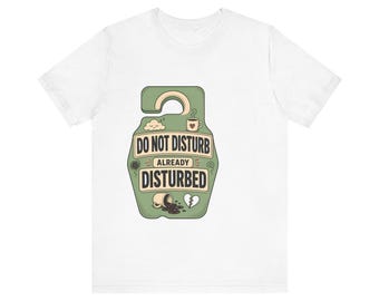 May include: A white short-sleeve t-shirt featuring a sage green door hanger graphic. The design includes the text DO NOT DISTURB ALREADY DISTURBED, a sleeping cloud, a coffee mug with a heart, a spilled cup of coffee, and a broken heart icon.