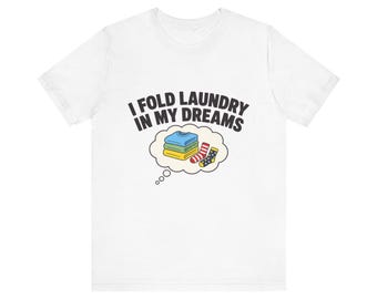 May include: A white short-sleeved t-shirt featuring the black text I FOLD LAUNDRY IN MY DREAMS above a thought bubble graphic. Inside the bubble is a stack of folded blue, green, and yellow shirts next to a red striped sock and a blue polka-dot sock.