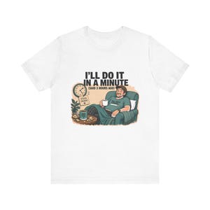 May include: A white t-shirt featuring a graphic of a person relaxing in a green armchair with a mug. The text reads I'LL DO IT IN A MINUTE (SAID 3 HOURS AGO), STUCK (AGAIN), DAD JOKES LOADING, and DAD FUEL. A clock and a small plant are also visible.