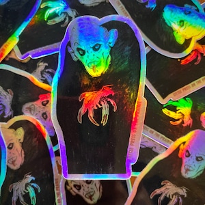 Puede incluir: Una pegatina holográfica con un vampiro con garras y colmillos afilados, con el texto "Little Press on the Prairie" en rojo.