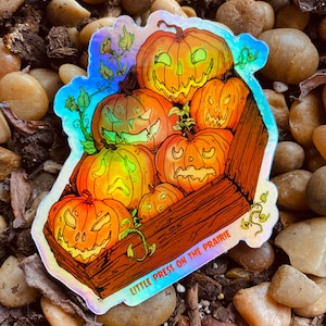 May include: A holographic sticker featuring a wooden crate overflowing with carved jack-o'-lantern pumpkins. The sticker has the text "Little Press on the Prairie" at the bottom.