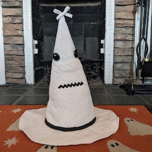 May include: A white quilted fabric cone-shaped hat with a wide brim, featuring two black circular button eyes, a black zigzag stitched mouth, and a small white fabric bow at the pointed top. The base of the cone is trimmed with a black band.