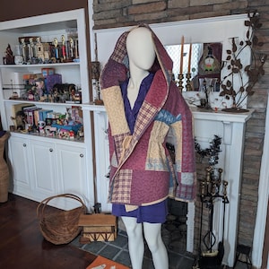 May include: A hooded jacket featuring a patchwork design with quilted textures in shades of maroon, tan, and blue. The garment has long sleeves, a drawstring waist, and is worn over a solid purple shirt. The fabric displays various patterns including plaid and swirling stitch details throughout the exterior.