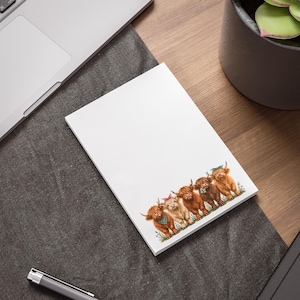 May include: A rectangular white notepad featuring a colorful illustration of five Highland cattle at the bottom. The cattle are depicted in shades of brown and cream, with some wearing small patterned bandanas or headbands in green and blue.