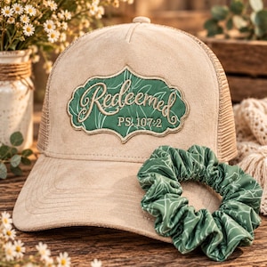 May include: A beige trucker hat with a mesh back and a suede-like front panel featuring a green patch with the embroidered text "Redeemed PS. 107:2" in a white script font. A matching green scrunchie with a white leaf pattern rests on the brim.