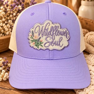 May include: A lavender baseball cap with a beige mesh back. The front features an embroidered patch with the text Wildflower Soul in purple script, accompanied by a small floral design with pink flowers and green stems on a cream background.