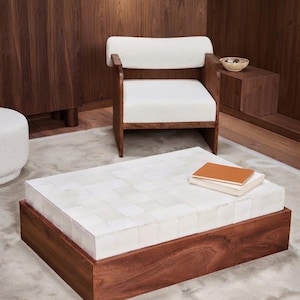 May include: A low rectangular coffee table featuring a dark wood base and a top surface composed of a grid of square white tiles. Two stacked books, one with a tan cover and one with a white cover, rest on the tiled surface of the table.