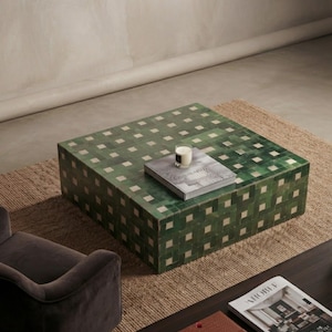 Pode incluir: Uma mesa de centro quadrada com um padrão geométrico de grelha de azulejos alternados em verde escuro e branco sujo. A superfície da mesa contém um livro cinzento com uma pequena vela de vidro por cima. O design cobre o topo e os quatro lados.