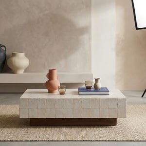 May include: A rectangular coffee table featuring a white tiled surface and a dark wooden base. On the table sits a terracotta-coloured vase, two small glass tumblers, a blue book, and a small off-white vase. The table is placed on a woven rug.