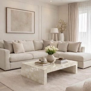 May include: A large beige sectional sofa with multiple matching throw pillows sits on a light-coloured rug. In front is a low, rectangular coffee table covered in small, glossy, off-white square tiles. A vase of white tulips rests on the table.