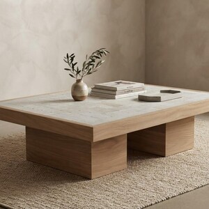 May include: A rectangular coffee table with a light wood frame and a textured, light grey stone tile surface. The table features two block-style wooden legs and supports a small ceramic vase with green leaves, a stack of books, and a tray.