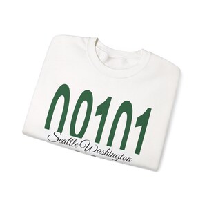 Könnte beinhalten: Ein weißes Sweatshirt mit Rundhalsausschnitt, das den dunkelgrünen Text 00101 und den schwarzen Schreibschrifttext Seattle Washington auf der Brust zeigt. Das Kleidungsstück hat lange Ärmel, einen gerippten Kragen und einen klassischen Schnitt.