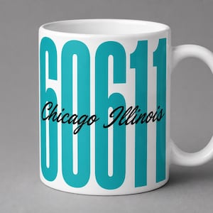 May include: A white ceramic mug featuring the large teal numbers 60611 printed vertically across the front. Overlaid across the center of the numbers is the text Chicago Illinois written in a black cursive script font with a handle on the right.