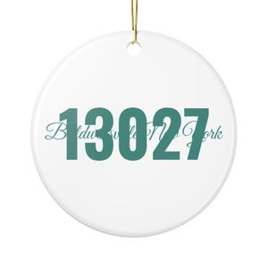 May include: A round white ceramic ornament featuring the teal text 13027 in a bold sans-serif font, with the teal cursive script text Baldwinsville New York layered behind it. A gold string is attached to the top for hanging purposes.