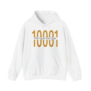 Puede incluir: Una sudadera con capucha blanca que presenta un gran gráfico dorado del número 10001 impreso dos veces, con el texto New York New York en una fuente cursiva negra posicionado horizontalmente entre los dos números en el pecho.