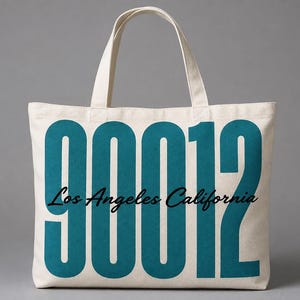May include: A cream-coloured canvas tote bag featuring large teal numbers 90012 printed on the front. Overlaid across the centre of the numbers is the text Los Angeles California in a black cursive script font. The bag has two matching fabric handles.