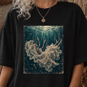 Puede incluir: Una camiseta negra con un estampado gráfico de un esqueleto con vestido de novia y velo de encaje blanco, flotando bajo el agua entre burbujas de aire. La escena se sitúa sobre un fondo acuático azul oscuro con rayos de luz.