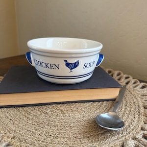 May include: A white ceramic soup bowl with two handles, featuring blue text that reads CHICKEN SOUP and a blue graphic of a chicken. The bowl has two horizontal blue lines circling the base and is accompanied by a small metal spoon.