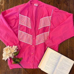 May include: A bright pink long-sleeve cardigan featuring a V-neckline and a front button closure. The garment includes two chevron-shaped panels of white and pink gingham fabric on the chest, accented with small floral embroidery and white piping.