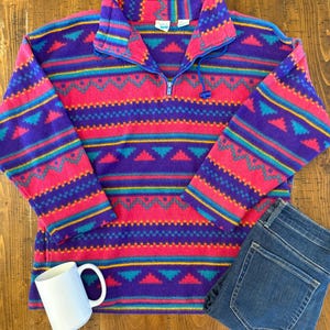May include: A long-sleeve fleece pullover featuring a vibrant geometric pattern in shades of purple, hot pink, teal, and yellow. The garment has a high collar with a quarter-length zipper closure, long sleeves, and a straight hemline.