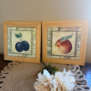 May include: Two square wooden frames containing artwork of fruit. The left frame displays two dark blue plums with green leaves. The right frame shows a single peach with a green leaf. Both prints feature a background with script text and a textured border.