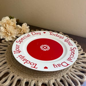 May include: A white ceramic plate with a wide red center circle featuring a small white floral wreath design. The rim of the plate is white with the red text Today's Your Special Day repeated twice, separated by two small red heart icons.