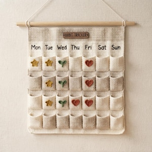 May include: A beige crocheted wall hanging habit tracker with a wooden rod and a label reading HABIT TRACKER. It features seven columns labeled Mon through Sun and four rows of pockets, decorated with small crocheted stars, leaves, and hearts.