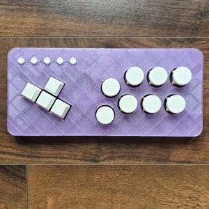 Pode incluir: Um comando de jogo retangular roxo com superfície texturizada, cinco pequenos botões circulares brancos em linha, quatro teclas quadradas brancas num grupo direcional e seis botões circulares brancos maiores no lado direito.