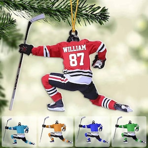 May include: A hockey player ornament featuring a red jersey with the name WILLIAM and number 87 on the back, black pants, and skates. The figure holds a hockey stick in one hand. Additional versions in teal, orange, blue, and green are shown below.