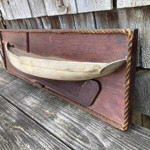 May include: A rectangular wooden wall plaque featuring a three-dimensional light-colored wooden boat model mounted on a dark brown wooden board. The plaque is framed with a thick, twisted tan rope border and includes a dark brown wooden rudder piece.