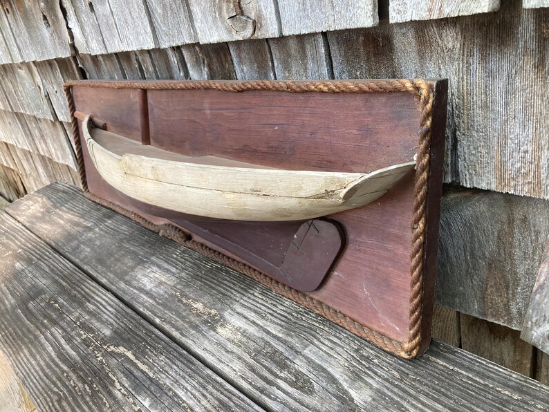 May include: A rectangular wooden wall plaque featuring a three-dimensional light-colored wooden boat model mounted on a dark brown wooden board. The plaque is framed with a thick, twisted tan rope border and includes a dark brown wooden rudder piece.