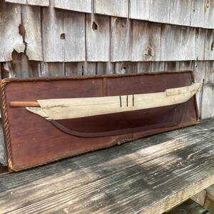 May include: A wall-mounted wooden plaque featuring a half-hull model of a boat. The plaque is rectangular with a rope border. The boat model has a light-colored upper hull with three vertical black lines and a dark brown lower hull section.