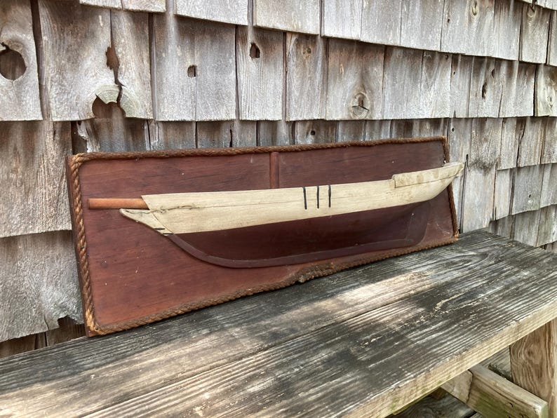 May include: A wall-mounted wooden plaque featuring a half-hull model of a boat. The plaque is rectangular with a rope border. The boat model has a light-colored upper hull with three vertical black lines and a dark brown lower hull section.