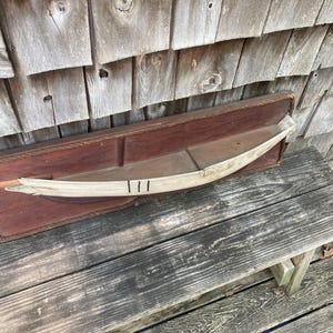 May include: A rectangular dark brown wooden wall plaque featuring a decorative rope border and a central horizontal shelf. Mounted on the shelf is a curved, light-colored wooden boat model with three distinct black vertical lines painted on it.