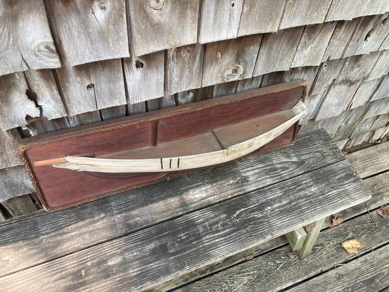 May include: A rectangular dark brown wooden wall plaque featuring a decorative rope border and a central horizontal shelf. Mounted on the shelf is a curved, light-colored wooden boat model with three distinct black vertical lines painted on it.