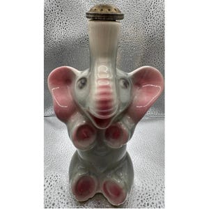 Peut inclure: Une carafe en céramique en forme d'éléphant avec une finition grise brillante et des accents roses sur les oreilles, les pieds et la trompe. La trompe relevée forme le goulot, surmonté d'un petit bouchon rond et texturé de couleur brune.