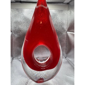 May include: A teardrop-shaped glass sculpture featuring a vibrant red core encased in clear glass. The piece has a prominent circular opening in the center and a textured, bubble-like pattern visible within the lower clear glass section.