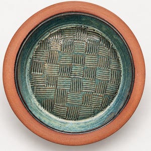 May include: A round ceramic bowl with a smooth, unglazed terracotta-colored rim and a deep teal interior. The center of the bowl features a raised, textured basket-weave pattern in a dark, mottled green glaze finish.