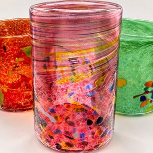 May include: Three cylindrical glass tumblers featuring speckled patterns. The center glass is pink with horizontal swirls, the left glass is orange with red and yellow flecks, and the right glass is green with multicolored spots.