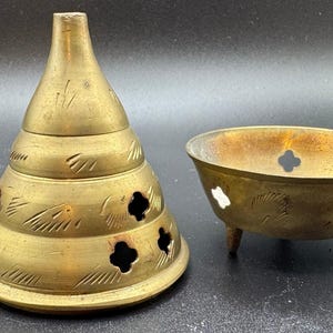 May include: A two-piece brass incense burner consisting of a conical lid and a matching bowl. The lid features a tiered design with small clover-shaped cutouts and etched markings. The bowl is supported by three small legs and has cutouts.