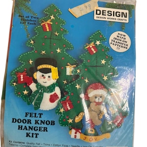 Puede incluir: Un kit de manualidades de fieltro para colgar en pomos de puertas con dos árboles de Navidad verdes. Un árbol incluye un muñeco de nieve con sombrero de copa negro, el otro un oso de peluche con gorro de Papá Noel. Texto en el paquete: Design Works Crafts, Set of Two Finishes, Now With Iron-On Transfer Patterns.