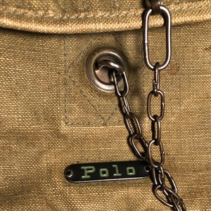 May include: A close-up of a brown canvas bag with a metal chain and a metal tag that says "Polo".