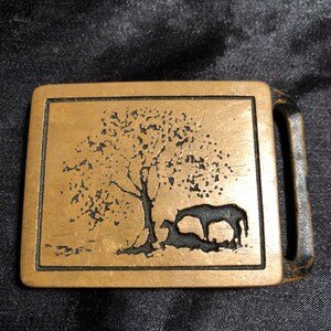 May include: A gold-toned metal belt buckle with a design of a tree and two horses. The buckle is rectangular with a slightly raised border.
