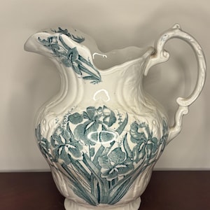 May include: A cream-colored ceramic pitcher featuring a teal-colored transferware pattern of iris flowers and foliage. The pitcher has a scalloped rim, a curved handle, and a molded relief texture on the body that complements the floral design.