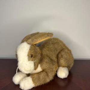 May include: A plush rabbit toy featuring brown and white fur. The rabbit has a white face, chest, and paws, with brown fur covering the rest of its body. It has long, upright ears, dark eyes, and thin white whiskers extending from its snout.