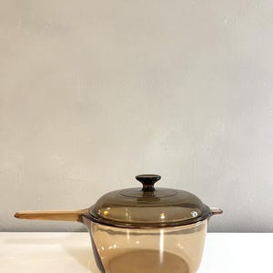May include: A translucent amber-colored glass saucepan with a matching glass lid. The pot features a long, integrated glass handle on one side and a small glass tab on the opposite side. The lid has a central knob handle for easy removal.