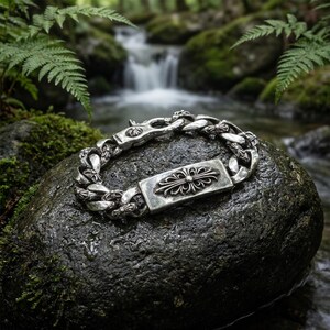 Puede incluir: Una pulsera de eslabones de plata con una placa de identificación rectangular que presenta un motivo de cruz floral en relieve. La pulsera incluye un cierre de mosquetón y detalles de cruces integrados en los eslabones, con acabado pulido.